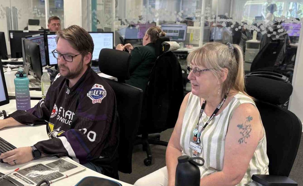 Image of Senior Engagement Officer Sue sat at a desk with someone from the Northwest Ambulance Service Emergency Operations Call Centre watching the process