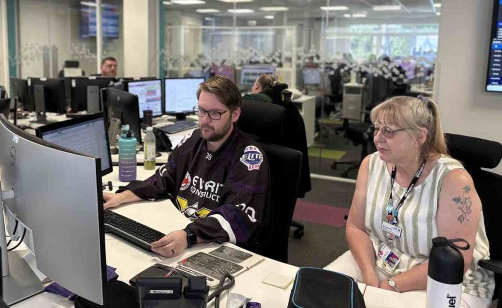Image of Senior Engagement Officer Sue sat at a desk with someone from the Northwest Ambulance Service Emergency Operations Call Centre watching the process