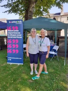 Image of Senior Engagement Officer Sue with a representative from ST John's Hospice at Bay Health Festival in Lancaster stoff infront of a navy blue Healthwatch Banner detailing our services.